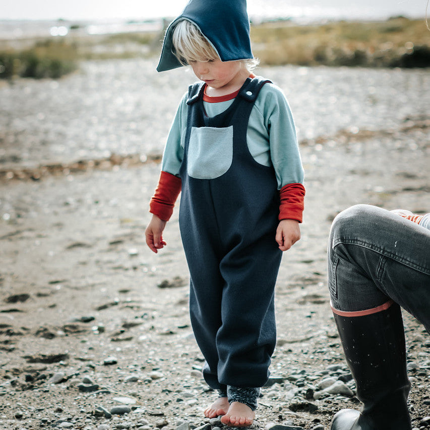 Child in blue outfit and green hat standing on a rocky shore with a person partially visible.