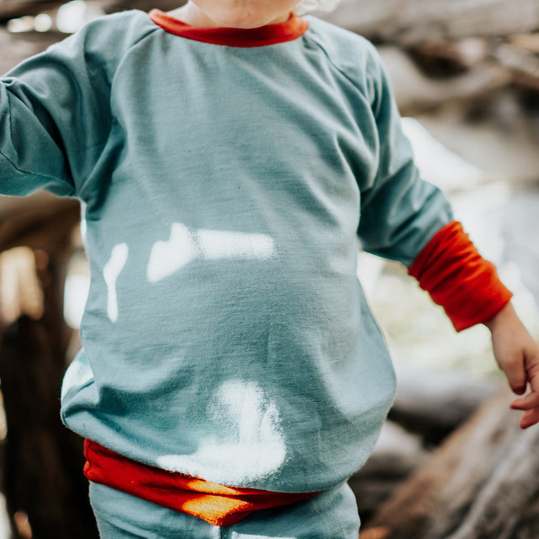 Child wearing a teal shirt with orange cuffs and pants, standing among wooden logs.