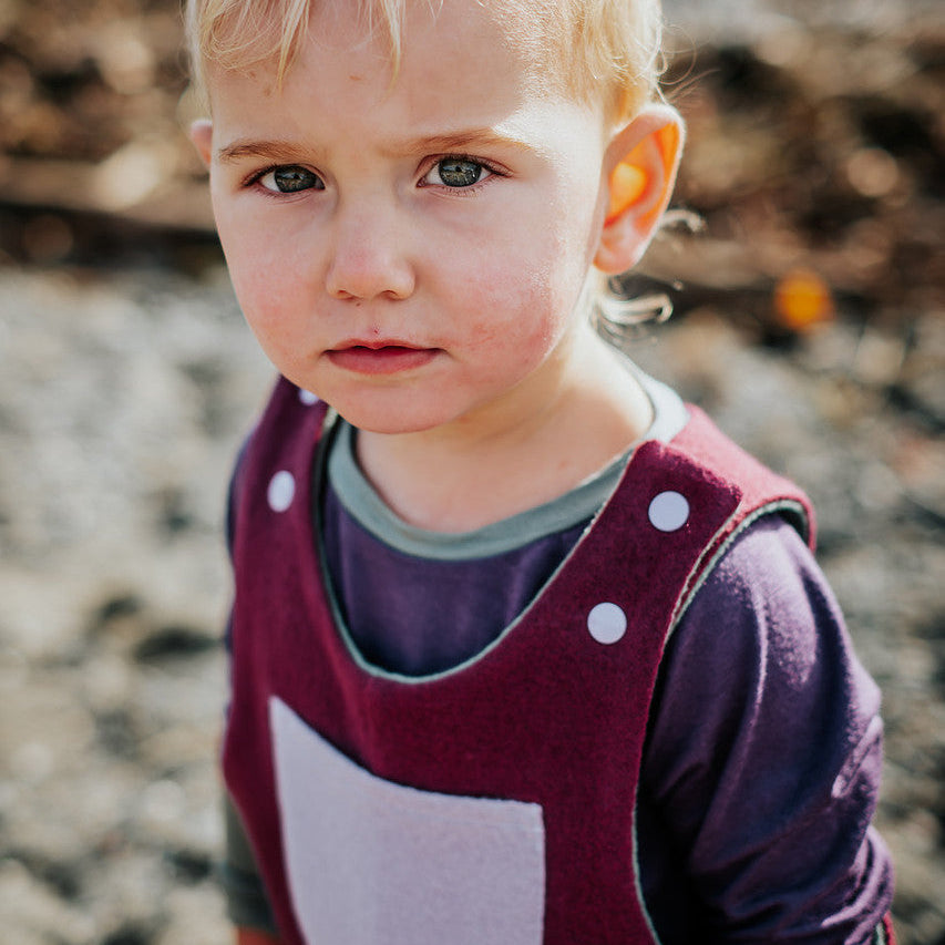 Young child wearing a maroon overalls with a white pocket, standing outdoors.