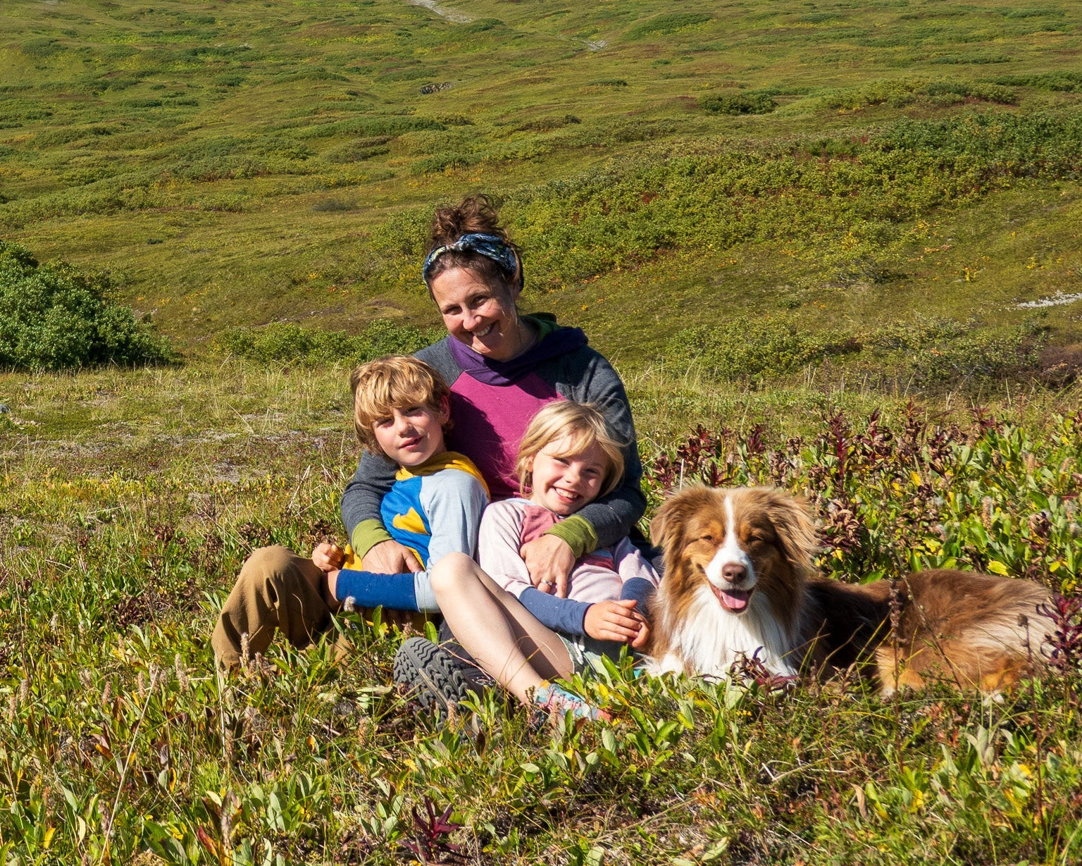 Woman with two children and a dog sitting in a grassy field with mountains in the background